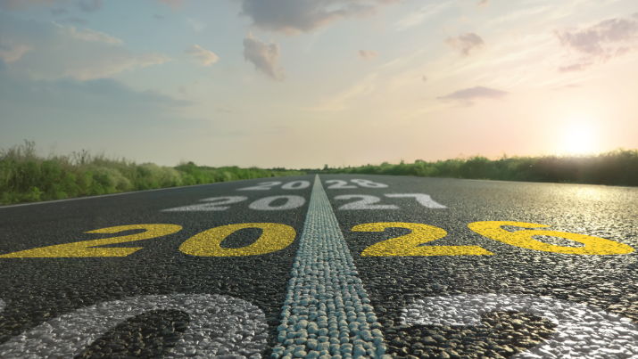 Auf einer Straße die bis zum Horizont führt stehen weiße Jahreszahlen geschrieben nur die 2026 ist in gelb.